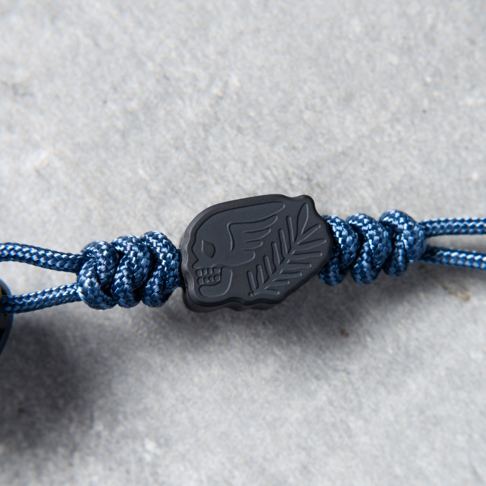 Close-up detail of the included lanyard attached to the Pro-Tech Avalon BSA26.Avalon.1, shown against a gray concrete surface. The paracord lanyard bead and cord are the focal point, with the base of the black aluminum handle visible at the top of the frame.