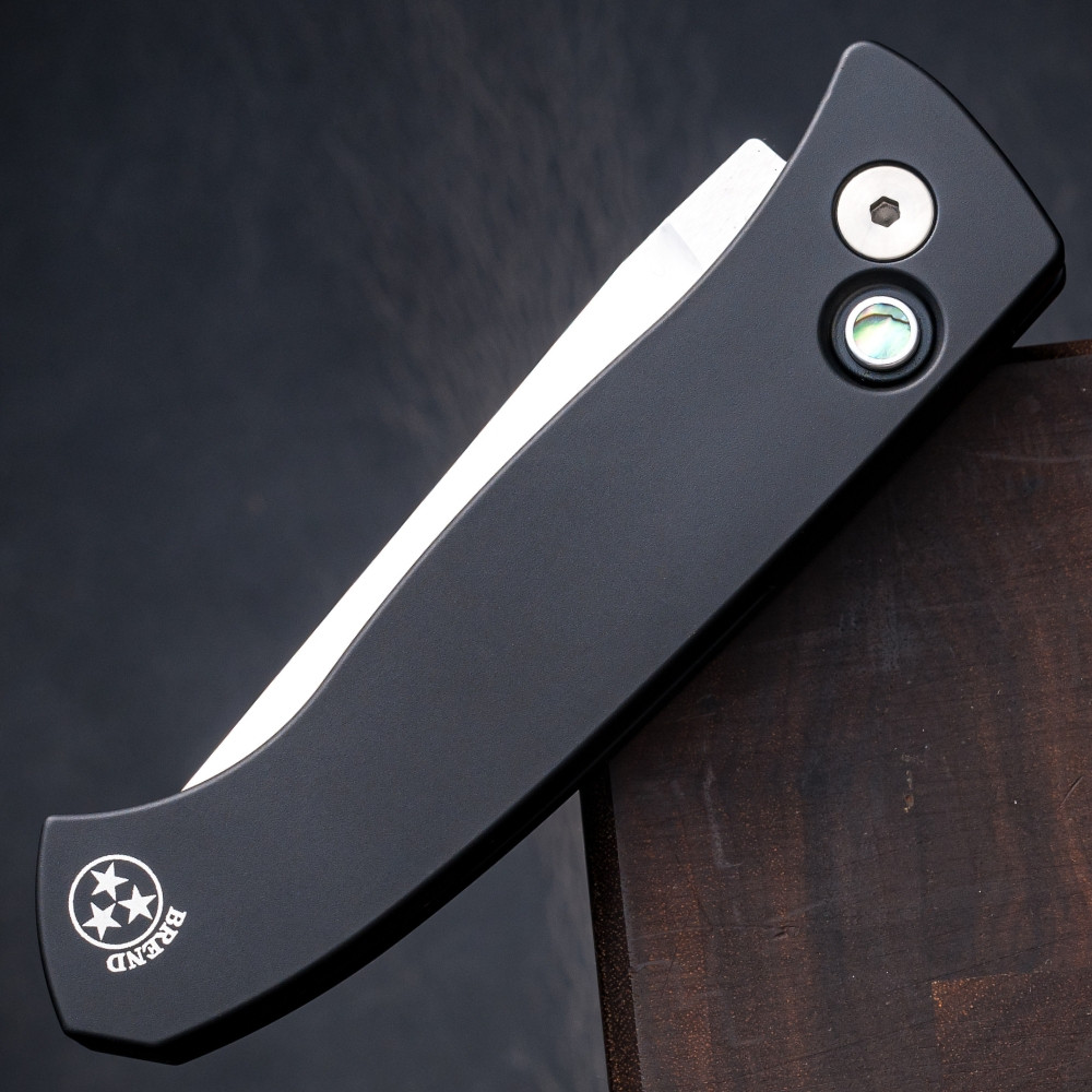 Pro-Tech Brend Classic BSA26.Brend.1 shown in a lengthwise profile against a dark walnut wood background, highlighting the knife's full silhouette in the closed or open position. The black aluminum handle, black pocket clip, and satin 154CM blade are visible from this angle.