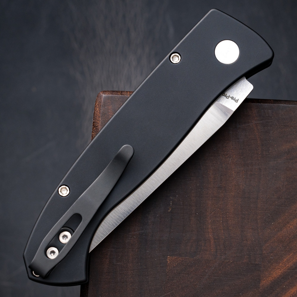 Close-up detail of the Pro-Tech Brend Classic BSA26.Brend.1 handle against a dark walnut wood background. The solid black aluminum scales, abalone button release, and satin hardware are the focal point of this shot.