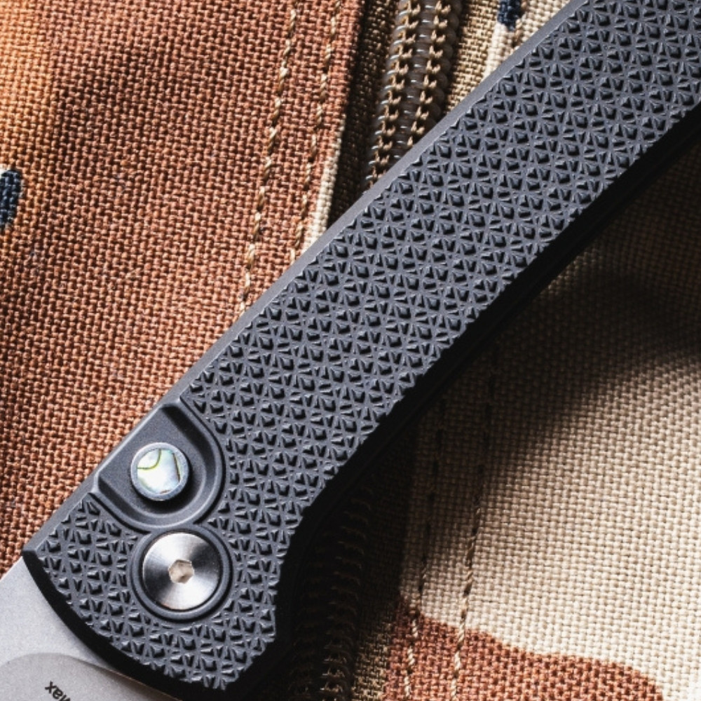 Close-up detail of the Pro-Tech Chaves Scapegoat Compact BSA26.Scapegoat.1 handle against the desert camouflage background, showing the aggressive black carbide diamond crosshatch texture in full detail. The round abalone push-button release and satin hardware are visible against the textured black aluminum scales.