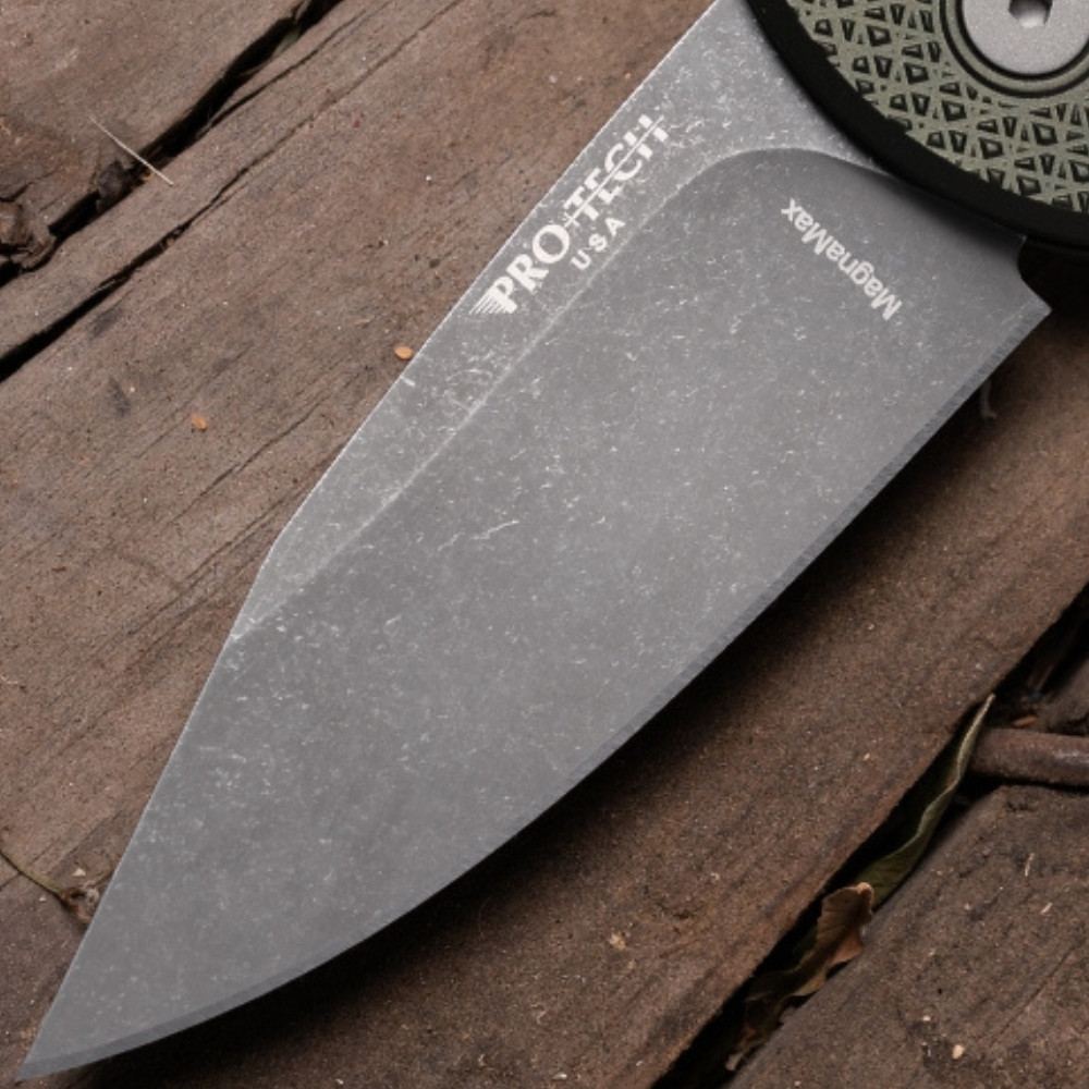 Close-up of the Pro-Tech Chaves Scapegoat Compact BSA26.Scapegoat.2 blade on a dark wood surface, showing the acid wash MagnaMax finish in detail. The layered, organic surface texture of the acid wash treatment and the clip point blade geometry are the focal point of this shot.