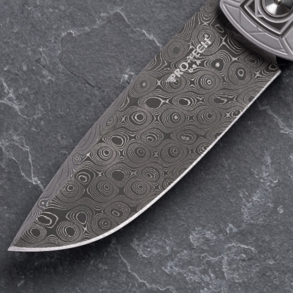 Close-up of the Pro-Tech VERO Synapse 2 Custom 002 Chad Nichols Raindrop Damascus blade on a gray slate surface. The distinctive concentric swirling raindrop pattern of the Damascus steel is the focal point, showing the full width and depth of the pattern across the blade face.
