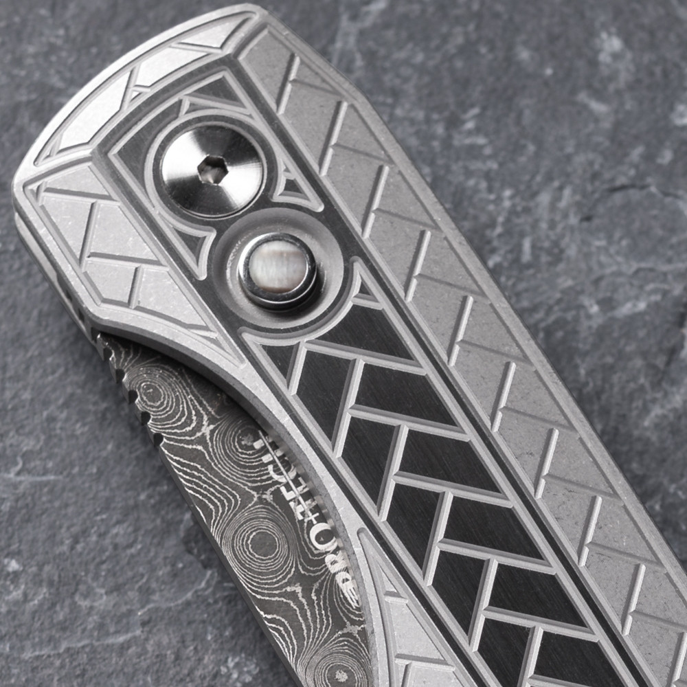 Close-up detail of the Pro-Tech VERO Synapse 2 Custom 002 handle on a gray slate surface. The deep herringbone milled texture and 2-tone blasted and tumbled finish of the 17-4 heat treated steel scales are shown in full detail, with the black lip pearl push-button release clearly visible against the textured handle.