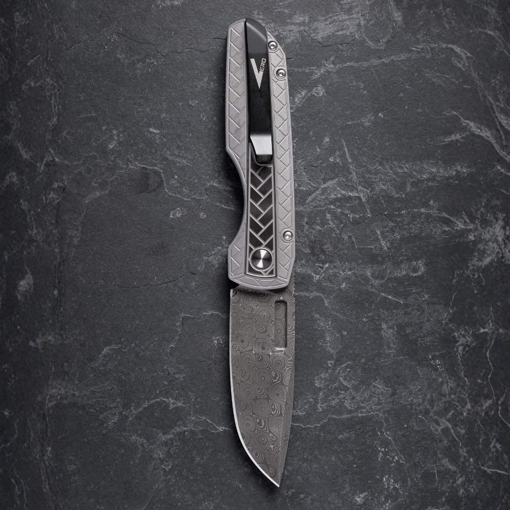 Pro-Tech VERO Synapse 2 Custom 002 shown from the clip side on a gray slate surface. The DLC machined titanium clip is prominently visible on the spine of the 17-4 herringbone steel handle, with the Raindrop Damascus blade partially visible at the base of the frame.