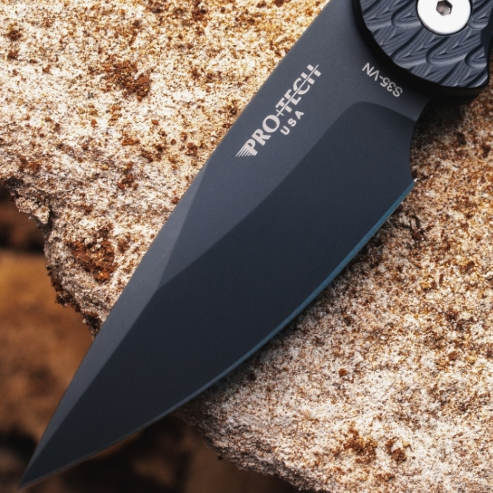 Pro-Tech TR-5 BSA26.TR5.1 shown open on a rough stone surface from a slightly different angle. The DLC black blade and feather-textured black aluminum handle are both visible, with the black lip pearl push button and black deep-carry clip in frame against the stone background.