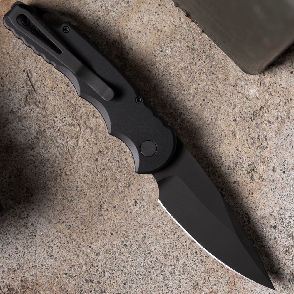 Pro-Tech TR-5 Operator T503-Operator shown open on a sandy concrete surface from a slightly elevated angle. The sterile DLC black blade is deployed, and the all-black aluminum handle, black hardware, and black deep-carry clip are fully visible with no identifying markings anywhere on the knife.