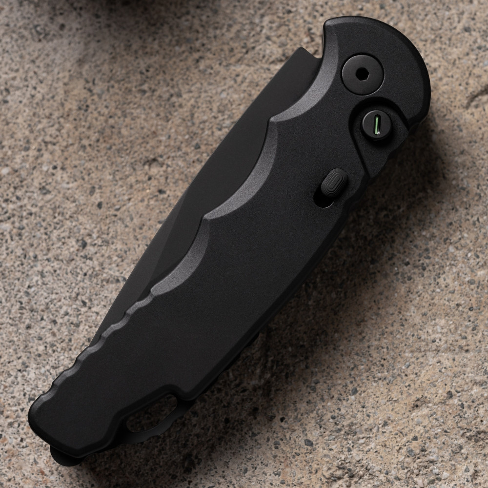 Pro-Tech TR-5 Operator T503-Operator shown at an angled profile on a sandy concrete surface, displaying the handle detail including the slide safety and tritium inlaid push button. The fully blacked-out DLC finish on the handle and hardware is the focal point of this close perspective.
