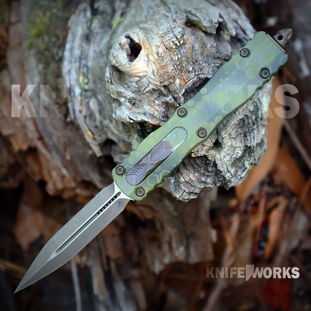 The Microtech Dirac OTF (225-13APBOHXCS) shown with its 2.92-inch Böhler M390MK double-edge dagger blade in a Bronzed Apocalyptic finish, Bazooka Hex Camo 6061-T6 aluminum handle with bronze hardware, and its tip-down right/left carry pocket clip. The front thumb slider and glass breaker are visible on this OTF automatic. ⚠️ This is both an OTF automatic and a double-edged blade — verify legal status in your jurisdiction before purchasing.