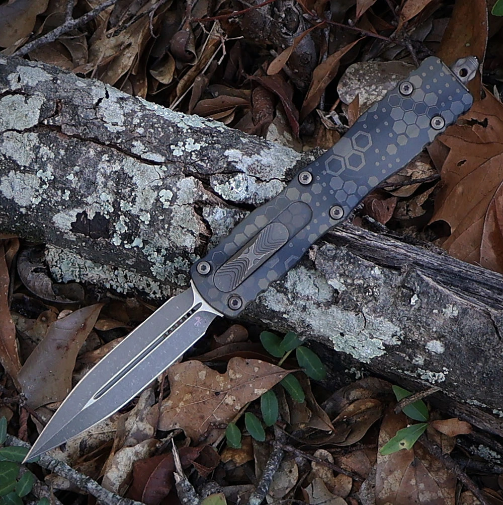 The Microtech Dirac Delta OTF knife (227-13 APBNHXCS) is shown with the blade extended against a neutral background. The double edge dagger-style blade has a bronze-toned Bronzed Apocalyptic finish, deployed from a 6061-T6 aluminum handle finished in a tan-and-brown Badlands Hex Camo pattern with a side button visible along the handle spine.