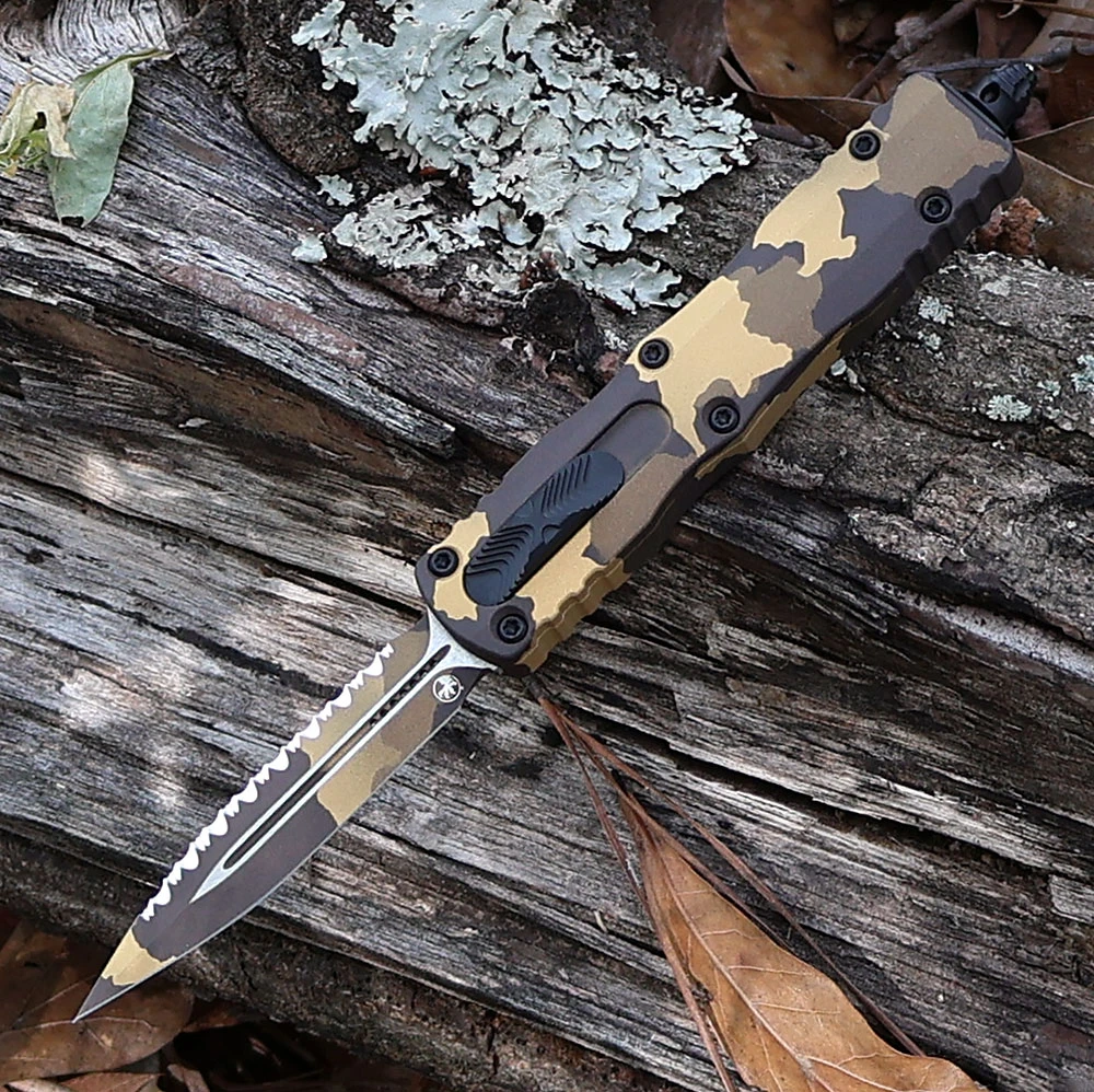 A Microtech Dirac Signature Series 225-3GCS-Z4 OTF automatic knife shown with blade deployed. The 2.92-inch double edge dagger blade is visible in Gold Camo finish with a flat grind; one edge is plain and one edge is serrated. The blade is 0.095 inches thin. The Gold Camo Mil-Spec Hard Coat anodized 6061-T6 aluminum handle shows matching Gold Camo finish at 0.42 inches thick. The cover-firing slider is visible on the handle for dual-action OTF deployment and retraction. The 301 stainless steel tip-down clip is mounted. Glass breaker visible at the handle end. Overall 7.25 inches, closed 4.33 inches, 2.2 oz. Tony Marfione design. Made in the USA. ⚠️ OTF automatic, double-edged — legal where permitted.