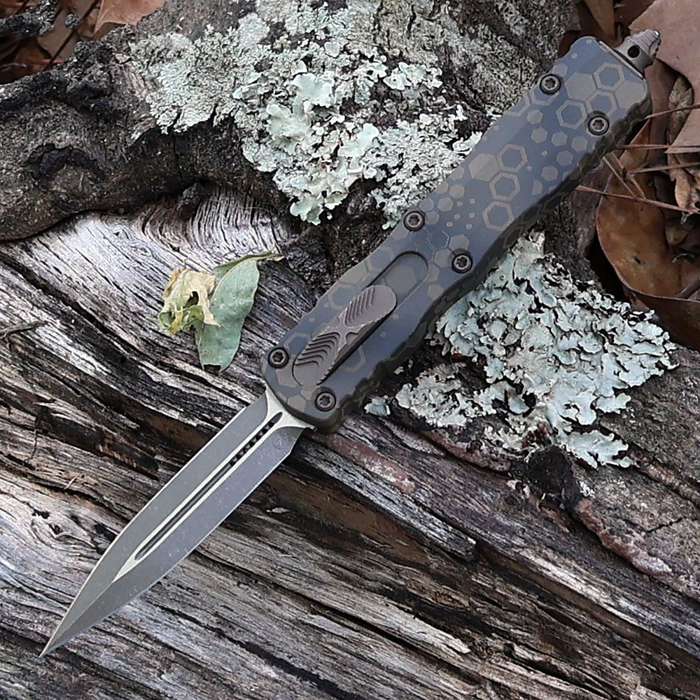 A Microtech Dirac Signature Series 225-13APBNHXCS OTF automatic knife shown with blade deployed. The 2.92-inch Böhler M390MK blade is visible in Bronzed Apocalyptic finish — a warm-toned mottled chemical treatment with bronzed character visible across the flat grind surface at 0.079 inches thin; the plain edge runs 2.64 inches of cutting edge. The Badlands Hex Camo aluminum handle displays a hexagonal patterned camo treatment in an earth-tone Badlands colorway at 0.44 inches thick. The cover-firing slider is visible for dual-action OTF deployment and retraction. Glass breaker visible at handle end. Tip-down clip mounted. Overall 7.25 inches, weight 2.19 oz. Tony Marfione design, Signature Series. Made in the USA. ⚠️ OTF automatic — legal where permitted.