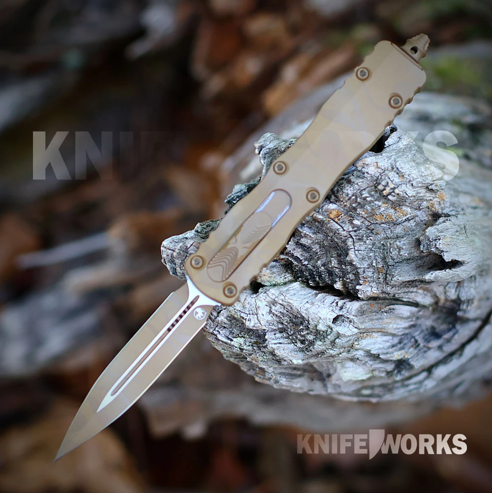 The Microtech Dirac D/E is shown with its double-edge dagger blade deployed from the front of the curved aluminum handle. The blade and handle both carry a Sand Dune Camo pattern, and the slider and pocket clip are visible.