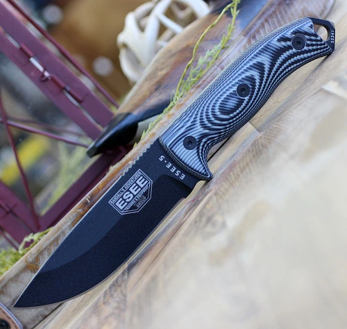 The ESEE-5 3D Handle (5PB-002) shown with its 5.25-inch 1095 saber ground drop point blade in a black finish, sharpened glass breaker pommel, fire-making bow drill divot, 3D gray and black G-10 handle scales, and its black Kydex belt sheath.