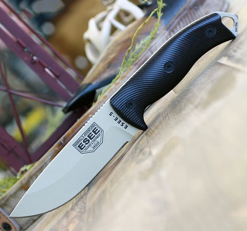The ESEE-5 3D Handle (5PDT-004) shown with its 5.25-inch 1095 saber ground drop point blade in a Desert Tan finish, sharpened glass breaker pommel, fire-making bow drill divot, 3D Blood and black G-10 handle scales, and its black Kydex belt sheath.