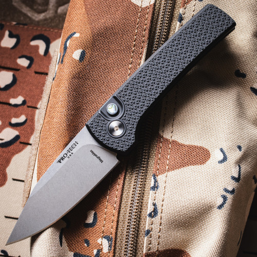 Pro-Tech Chaves Scapegoat Compact BSA26.Scapegoat.1 shown open on a desert camouflage bag background. The full knife is visible with the stonewash MagnaMax clip point blade deployed, black carbide-textured aluminum handle prominent, and the abalone push button and satin hardware visible on the frame.