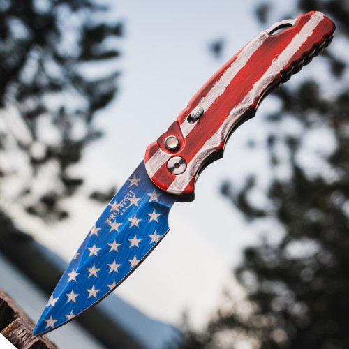 Pro-Tech TR-5 T501-PATRIOT shown deployed against an outdoor blue sky background. The USA flag Patriot Cerakote finish is clearly visible across both the handle and blade, with red and white stripes and blue field with white stars covering the full knife. The gold lip pearl push button and deep-carry pocket clip are visible on the handle.