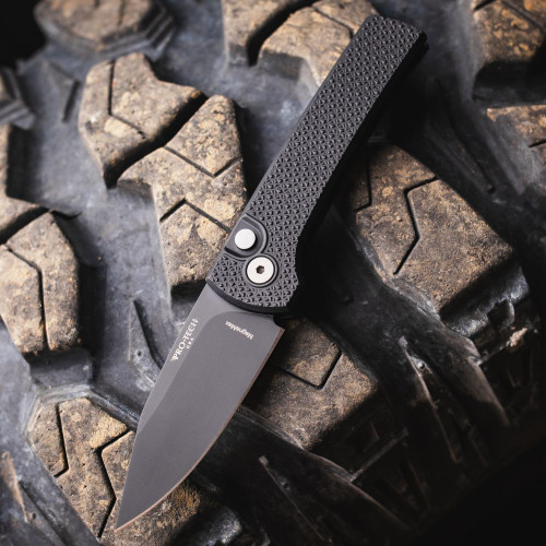 Pro-Tech Scapegoat Compact Auto RCS1106 — open, MagnaMax DLC black blade extended, carbide textured black aluminum handle, cracked earth background