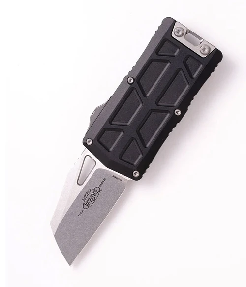 A Microtech Exocet® II 1156-10 double-action OTF money clip shown with blade deployed. The 2.34-inch Wharncliffe blade is visible in stonewash finish; the straight primary edge running parallel to the handle spine and the curved spine descending to a fine tip are clearly visible. The black handle frame functions as the money clip — shown at 0.39 inches thin and 1.48 inches wide. The double-action thumb slide for OTF deployment and retraction is visible on the handle. Overall length 5.69 inches. Made in the USA by Microtech Knives. ⚠️ OTF automatic — legal where permitted.