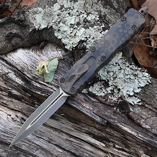 A Microtech Dirac Signature Series 225-13APBNHXCS OTF automatic knife shown with blade deployed. The 2.92-inch Böhler M390MK blade is visible in Bronzed Apocalyptic finish — a warm-toned mottled chemical treatment with bronzed character visible across the flat grind surface at 0.079 inches thin; the plain edge runs 2.64 inches of cutting edge. The Badlands Hex Camo aluminum handle displays a hexagonal patterned camo treatment in an earth-tone Badlands colorway at 0.44 inches thick. The cover-firing slider is visible for dual-action OTF deployment and retraction. Glass breaker visible at handle end. Tip-down clip mounted. Overall 7.25 inches, weight 2.19 oz. Tony Marfione design, Signature Series. Made in the USA. ⚠️ OTF automatic — legal where permitted.