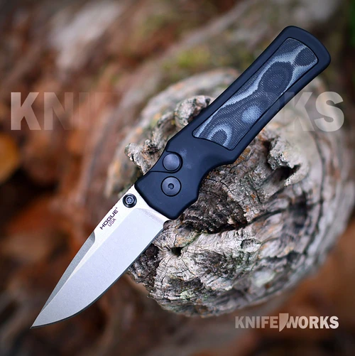 The Hogue Saxum 24679 is shown open on a weathered, lichen-covered log against a blurred autumn background. The stone tumbled drop point blade carries the Hogue USA logo and a black thumb stud, and the matte black aluminum handle holds a black and gray G-Mascus G10 insert behind a round push button.
