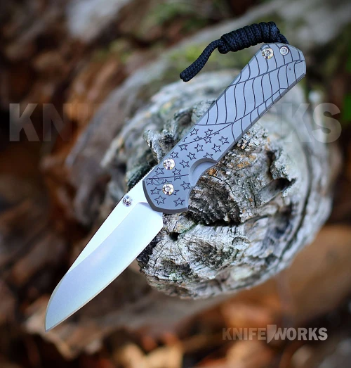 The Chris Reeve Small Sebenza 31 S31-1508 is shown open on a weathered log against a blurred autumn background. The Insingo blade extends from a glass blasted titanium handle machined with the Forever Flag CGG's stars and waving stripes, with gold-tone screws and a black cord lanyard at the butt.