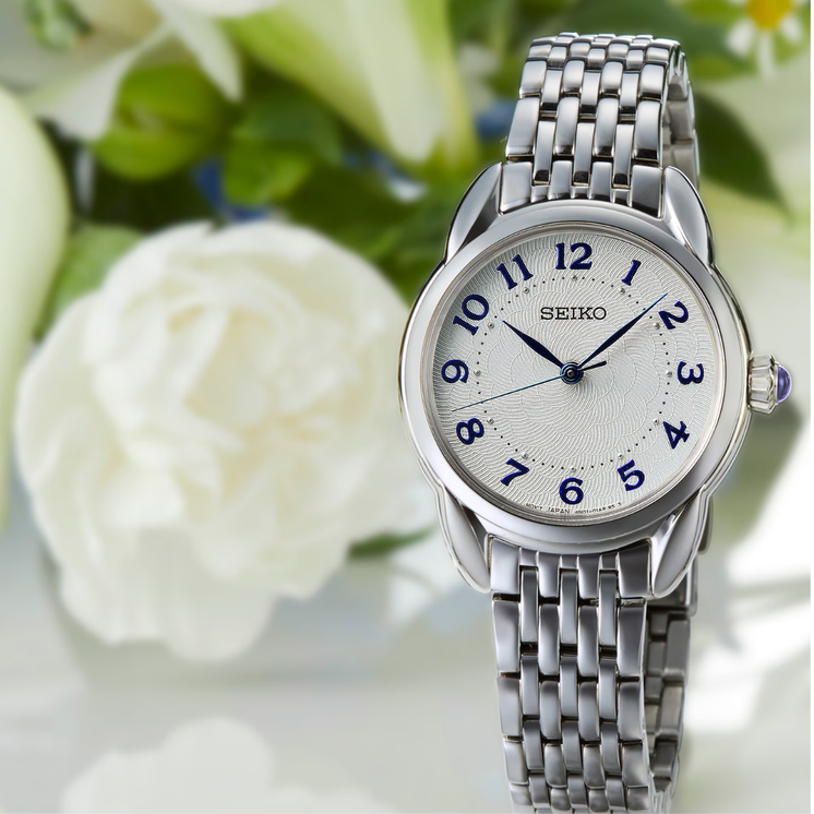 Seiko Ladies SUR561P1 con esfera blanca, números azules y brazalete de acero en composición lifestyle con flores blancas