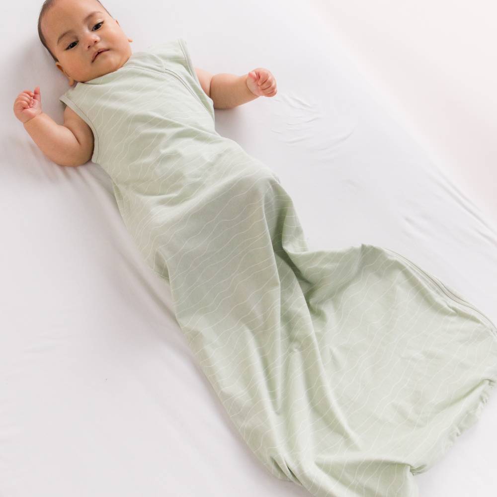 Baby lying in cot wearing the Baby Basics 0.2 tog cotton sleeping bag in avo wave print