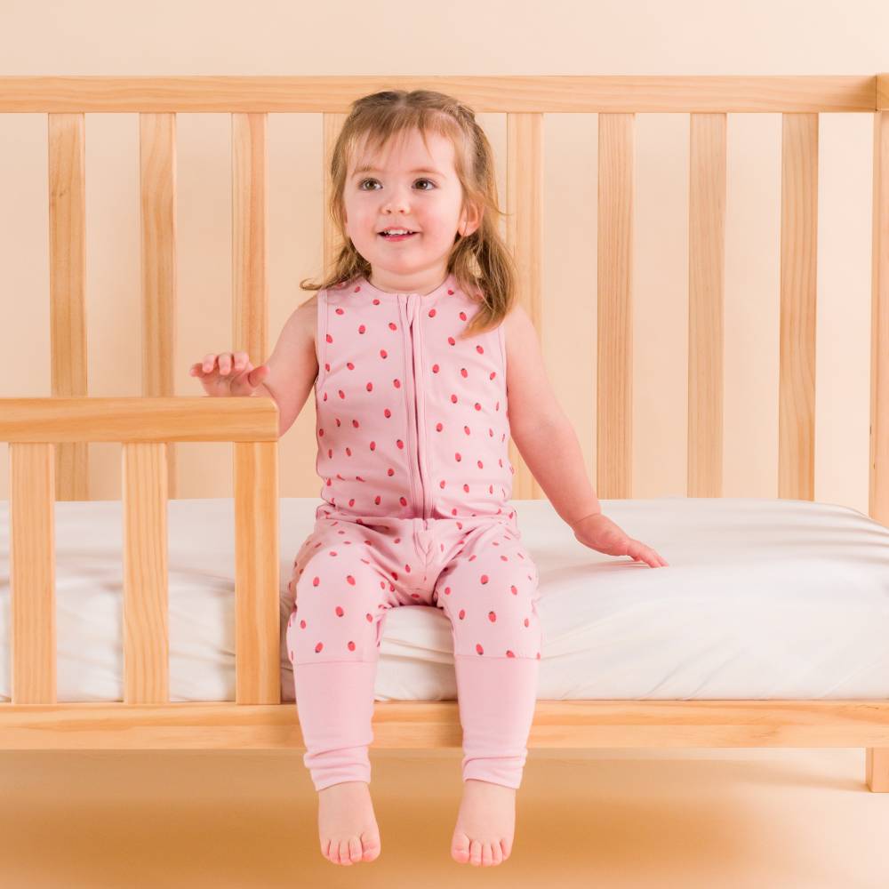 Girl sitting on side of cot wearing The Sleep Store Organic Cotton 0.2 tog Sleeping Suit in Blush strawberries