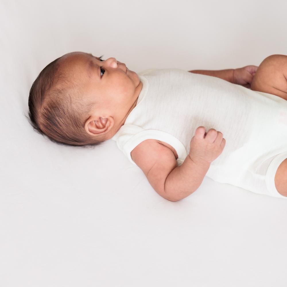 A baby lying down wearing The Sleeps Store's Basics Merino Singletsuit in the colour Cream.