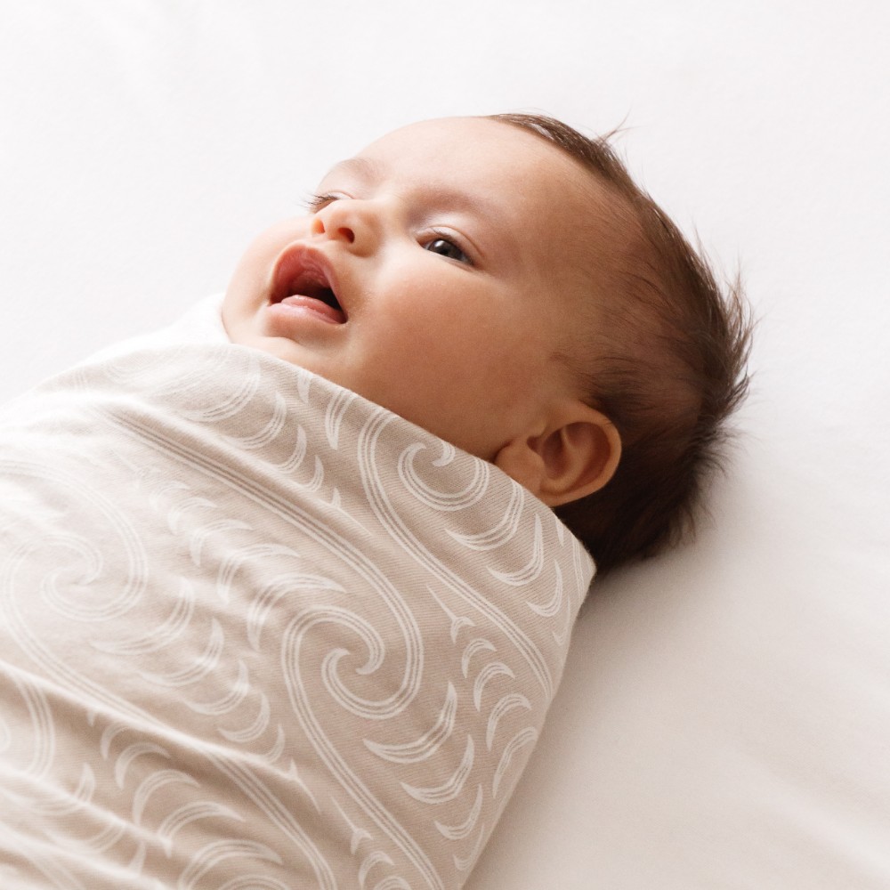 A baby lying down wrapped/swaddled in the Woolbabe X BO & KO Swaddle Blanket in Flax Ngaru.
