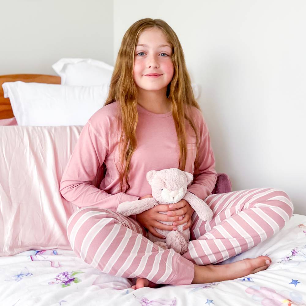 A girl sitting on her bed holding a teddy bear wearing the Woolbabe Merino/Organic Cotton Relaxed Winter Pyjamas in the colour Dusk/Shell.