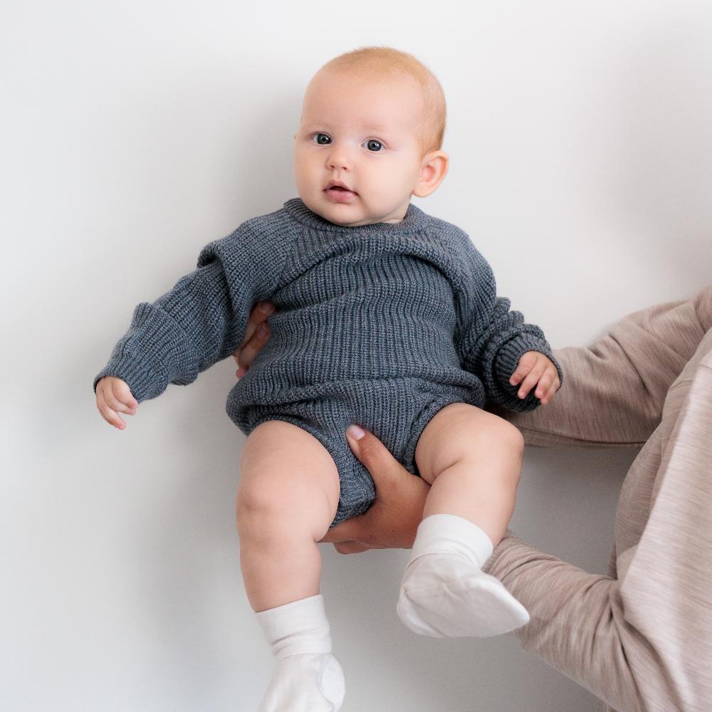 A baby being held front view wearing a Grey Marl Sleep Store Merino Knitwear Bubblesuit.