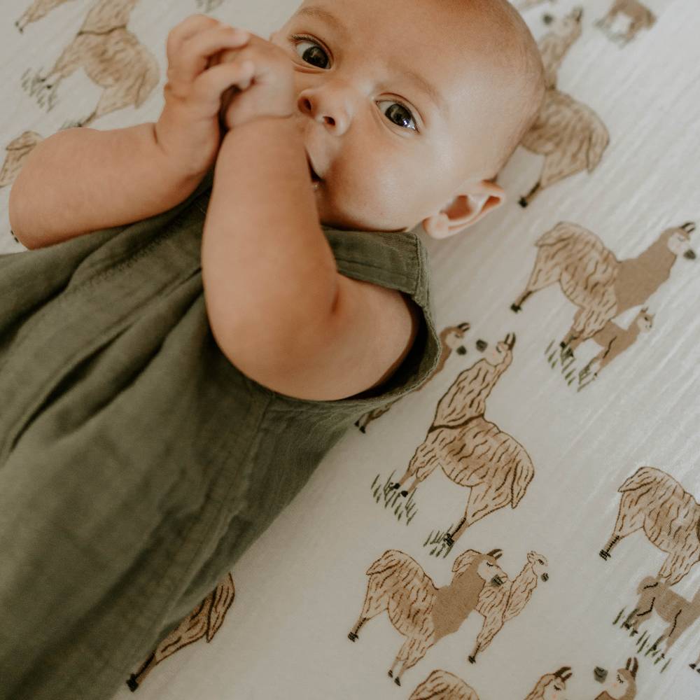 Baby sucking hands on Little Unicorn fitted cot sheet in Llama and wearing the Fern colour Little Unicorn Cotton Muslin Sleeping Bag