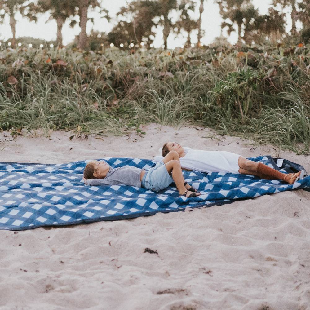 Two kids lying on Little Unicorn picnic blanket at the beach.