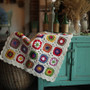 Multicolored handmade crochet daisy blanket with white trim on a teal cabinet with decorative items.