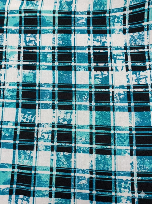 A top-down, flat lay photograph of a textile pattern. The fabric features a bold, grid-like plaid design composed of deep teal, turquoise, and black lines against a white background. The pattern has a distressed, textured appearance, with the lines showing speckled and broken effects, resembling worn paint or a hand-printed look.