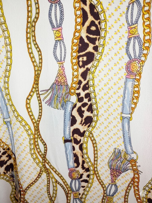 Extreme close-up of Poly Crepe Georgette fabric featuring a detailed equestrian scarf print with vibrant golden chains, multi-colored tassels, and bold leopard print stripes against a crisp ivory background.
