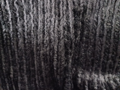 A detailed close-up shot of soft, plush, deep black velvet fabric. The material features distinct vertical ridges or stripes that add depth and texture to its velvety surface. Subtle lighting highlights the softness of the fibers.