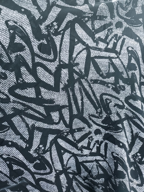 A close-up, high-angle shot of a Rayon Challis fabric featuring a bold, black abstract "scribble" or graffiti-style pattern. The background of the textile has a subtle blue-silver tinge rather than a flat white, highlighting the intricate weave and soft texture of the material. The fabric is slightly draped, showing realistic folds and shadows that emphasize its fluid movement.