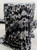 A Bolt of monochromatic abstract-print fabric elegantly draped over a pedestal against a bright, neutral light-gray studio background. The textile features a high-contrast pattern of swirling black, charcoal, and white tones, showcasing a soft, fluid drape and fine-knit texture.