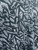 A close-up, high-angle shot of a Rayon Challis fabric featuring a bold, black abstract "scribble" or graffiti-style pattern. The background of the textile has a subtle blue-silver tinge rather than a flat white, highlighting the intricate weave and soft texture of the material. The fabric is slightly draped, showing realistic folds and shadows that emphasize its fluid movement.
