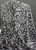 A close-up, studio-lit shot of Rayon Challis fabric featuring a bold black graffiti-style abstract pattern over a base with a subtle blue-silver tinge. The fabric is professionally draped to highlight its soft, fluid movement and lightweight texture, set against a seamless, simple gray background.