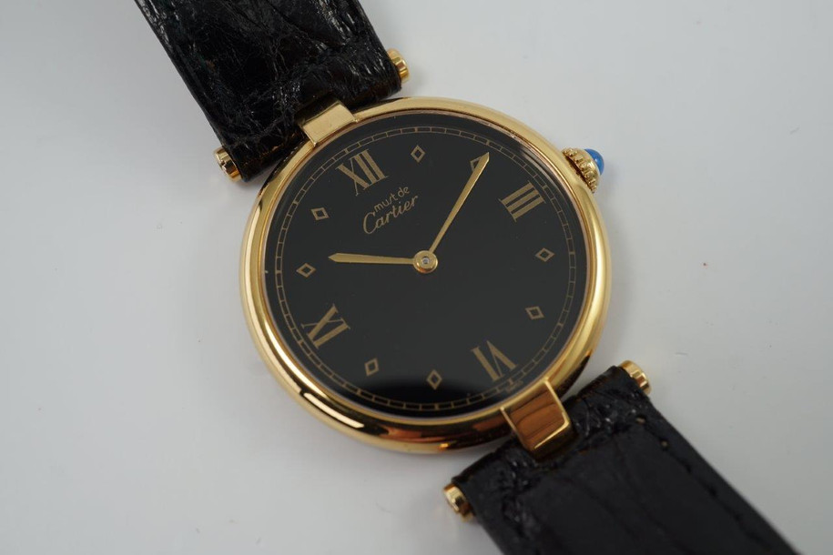 A very cool Cartier Vendome in vermeil, crafted during the 1990s. Believed to be new old stock for its condition, featuring gold diamond-shaped and Roman numeral hour markers, and gold applied sword hands and gold minute track all popping against the black dial with a light blue sapphire cabochon crown. The low-profile rounded case charmingly fastened by exposed t-bar lugs and a sumptuous leather strap with deployment buckle makes this piece comfortable to wear. Suitable for various occasions, for a man or woman. 

Original black dial, hands and Cartier sapphire crown. 
Cartier hang tag. 
Case measures 30 x 37mm, 5mm thick.
Silver stamps. 
Cartier Swiss caliber, quartz movement. 
Serial#5900xx 0669xx
Sapphire crystal.
Cartier black leather strap (appears in unworn condition, fits large and adjustable). 
Cartier gold plated deployant buckle. 
16mm lug width.
Modeled on 6 inch wrist.