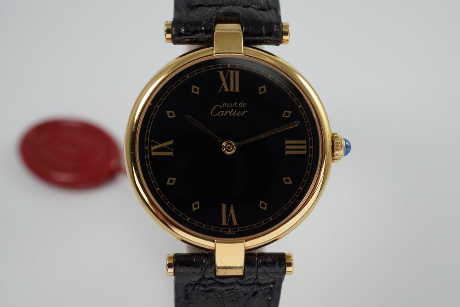 A very cool Cartier Vendome in vermeil, crafted during the 1990s. Believed to be new old stock for its condition, featuring gold diamond-shaped and Roman numeral hour markers, and gold applied sword hands and gold minute track all popping against the black dial with a light blue sapphire cabochon crown. The low-profile rounded case charmingly fastened by exposed t-bar lugs and a sumptuous leather strap with deployment buckle makes this piece comfortable to wear. Suitable for various occasions, for a man or woman. 

Original black dial, hands and Cartier sapphire crown. 
Cartier hang tag. 
Case measures 30 x 37mm, 5mm thick.
Silver stamps. 
Cartier Swiss caliber, quartz movement. 
Serial#5900xx 0669xx
Sapphire crystal.
Cartier black leather strap (appears in unworn condition, fits large and adjustable). 
Cartier gold plated deployant buckle. 
16mm lug width.
Modeled on 6 inch wrist.