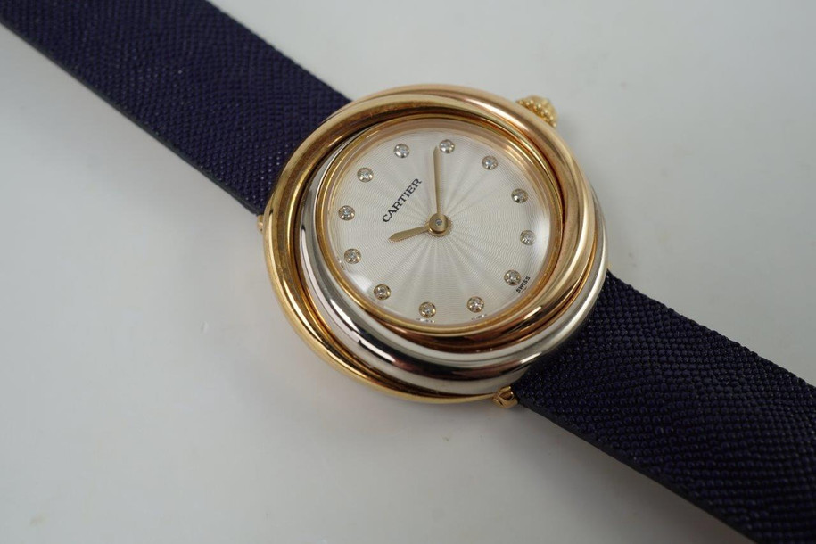 A fine preowned Cartier Trinity watch in 18k trio metals of white, yellow and rose gold, crafted circa 2000s. The trinity holds significance of fidelity, friendship, and love represented in the interlaced trio metals of the bezel, accented by a beaded diamond crown and guilloche dial with circular diamond hour markers and gold sword-shaped hands. The 26.5mm case holds presence on the wrist for its 7mm thick profile and is paired with a lovely shade of blue strap with hints of purple, accompanied by its original buckle. This accessory could take you from day to night, wearing beautifully in casual and formal settings.