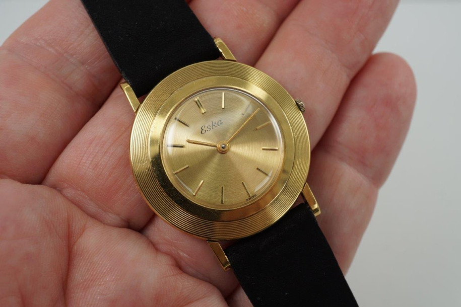 An Eska vintage watch in 18k yellow gold from the early 1960s. Regarded as a disco volante for its flying saucer-like appearance, the 33mm case holds a concentric reed-like textured bezel housing a gold-colored dial with gold stick hour markers and sword-shaped hands. Highly reflective qualities that would surely catch attention to the wearer’s wrist, the 7mm thick profile sits elegantly on the wrist from its overall delicate design. Inside of case back is signed S. Kocher Swiss, the name of Eska founders. Paired with a matte black soft suede-like strap, creates a striking contrast to the gold.