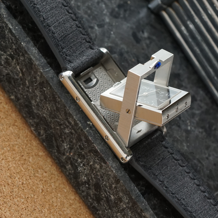 A late 1990s Cartier Tank Basculante in stainless steel, wonderfully constructed in a compact 22mm size. The dark grey patterned dial is contrasted by bright white stylized Arabic numerals and silvered sword-shaped hands, giving the watch a distinctive monochromatic look.
The Basculante's hinged case is the highlight of the design, allowing the watch head to pivot within its frame and turn completely over. It opens and closes smoothly and can remain upright within the frame. A blue cabochon accents the case, while the crown is cleverly concealed and accessed only when the watch is flipped from its usual position.
Powered by a quartz movement and fitted with a custom dark grey Babele leather strap secured by a Cartier stainless steel deployant clasp. An unusual interpretation of the Tank, with much of its appeal found in the ingenious case construction.

Watch Details
Reference: 2386
Case Number: 3514xxxx
Case: Stainless steel
Case Size: 22 × 32 mm; 6 mm thick
Dial: Dark grey patterned dial; white Arabic numerals
Movement: Quartz
Crystal: Sapphire
Strap: Custom dark grey Babele leather, adjustable
Lug Width: 17 mm
Clasp: Cartier stainless steel deployant
Wrist Size: Fits up to approximately 6 1/2 inches
Modelled On: 6-inch wrist
Condition: Light wear shown
Service: New battery installed