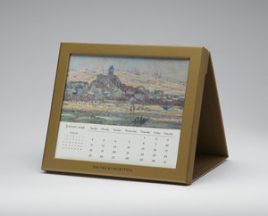 Desk calendar sitting on a white surface with an image of a painted cityscape by the water and the dates of the month of January 2026 below it, all framed by a golden border