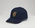 Navy baseball hat with the embroidered gold Frick Collection logo on the front sitting on a white surface.