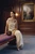 Model in a cream silk dress seated in an ornate wood-paneled room, with the rounded walnut wood Moon handbag resting beside her.